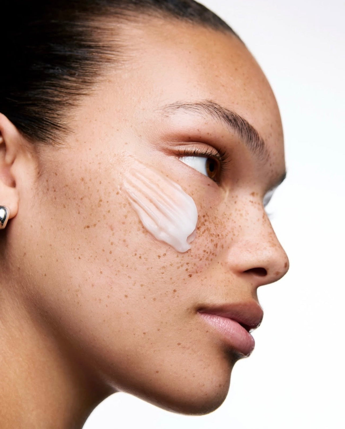 Close-up side profile of a woman with freckles applying or showing a white cream swatch on her cheek against a clean white background.